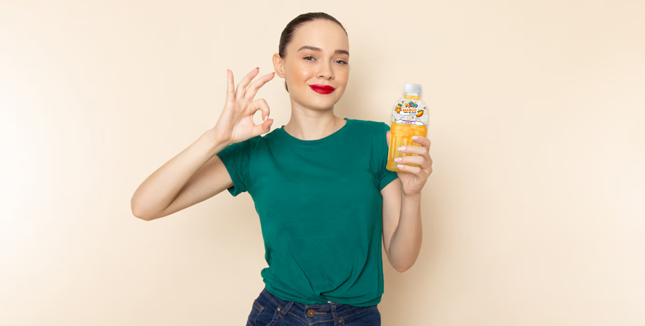 A woman in a green t-shirt holds a yellow bubble tea drink, making an "OK" hand gesture with a smile.